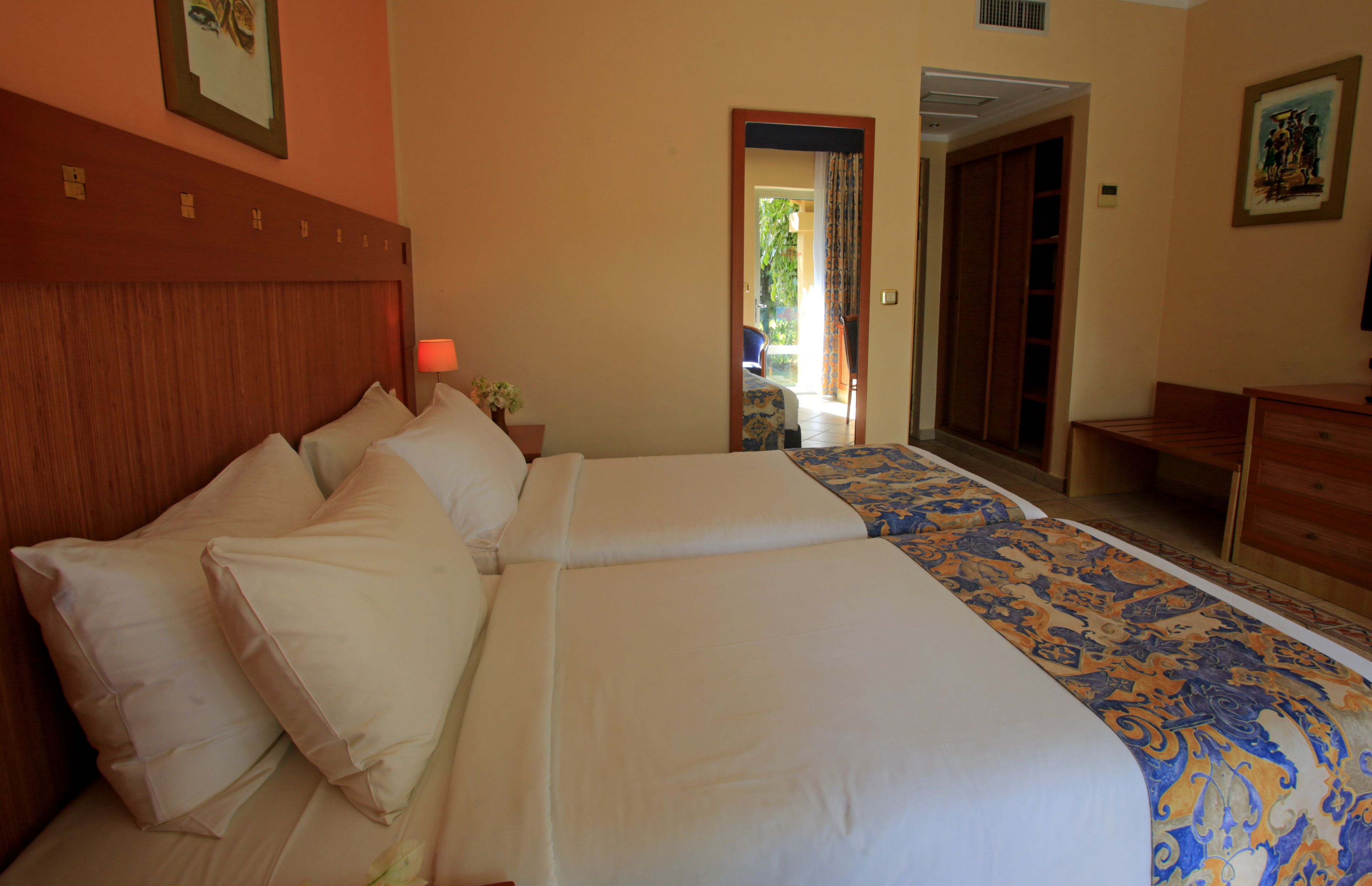 A hotel room with two beds, a wooden headboard, a nightstand with a lamp, and a door leading to a balcony.