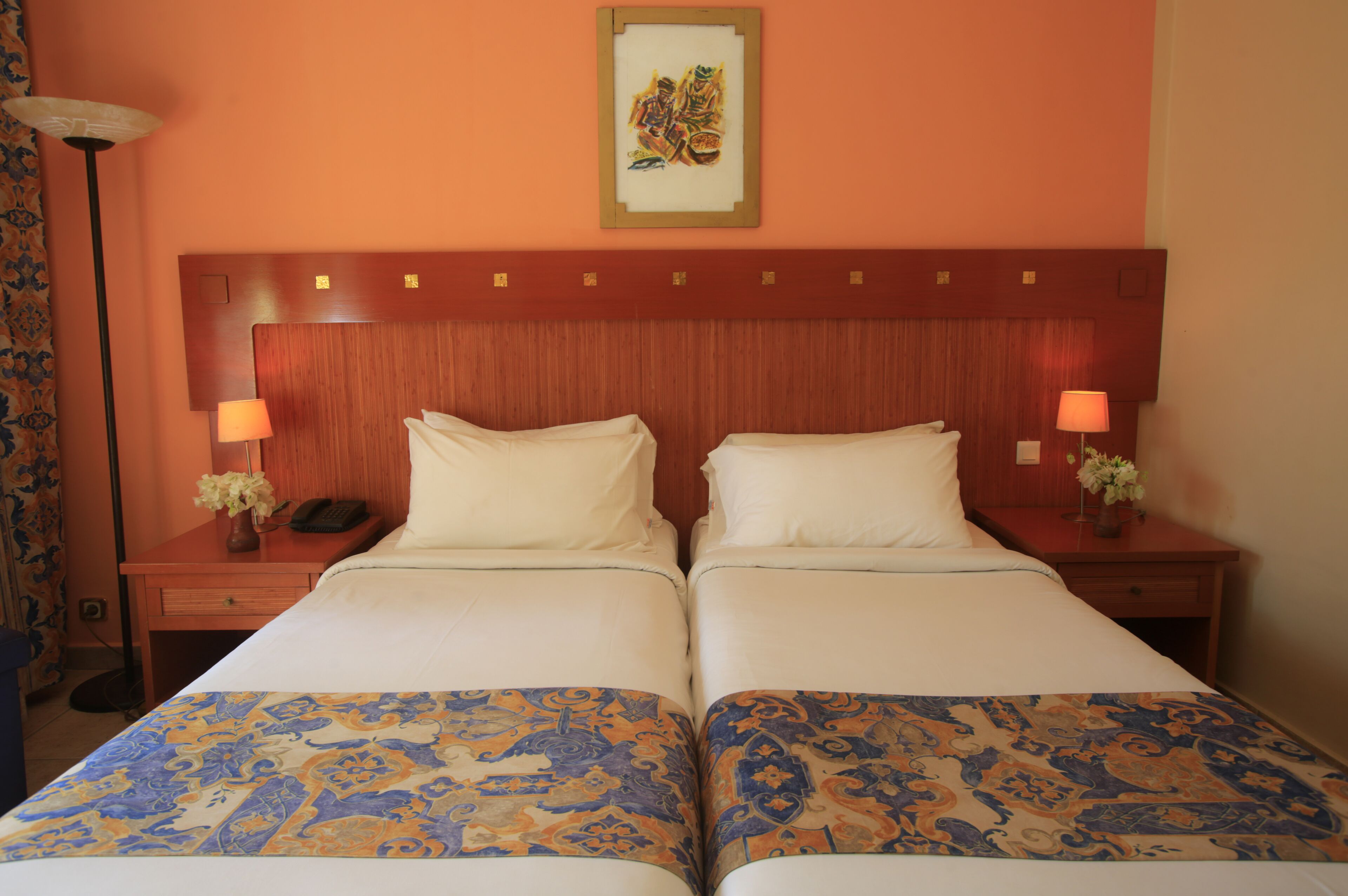 A double bed with a patterned bedspread, two bedside tables with lamps, a framed artwork above the bed, and a wooden headboard.