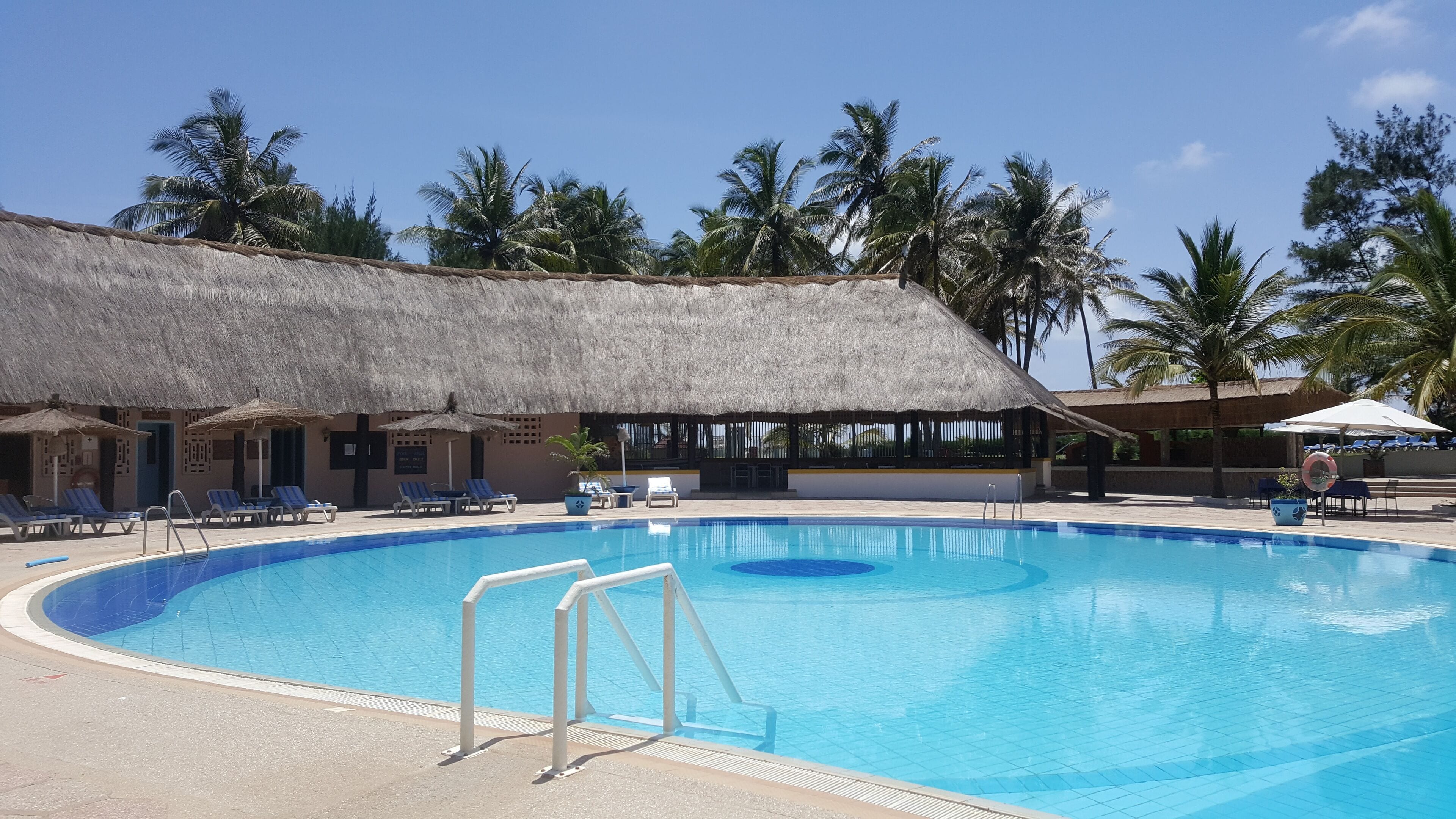 Piscina con sillas de descanso y sombrillas, rodeada de palmeras y un edificio con techo de paja.