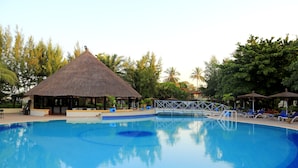 A swimming pool with lounge chairs and umbrellas, surrounded by trees and a thatched-roof structure. - The Kairaba Beach Hotel (Serrekunda)