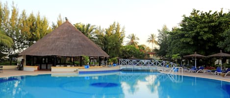 A swimming pool with lounge chairs and umbrellas, surrounded by trees and a thatched-roof structure.