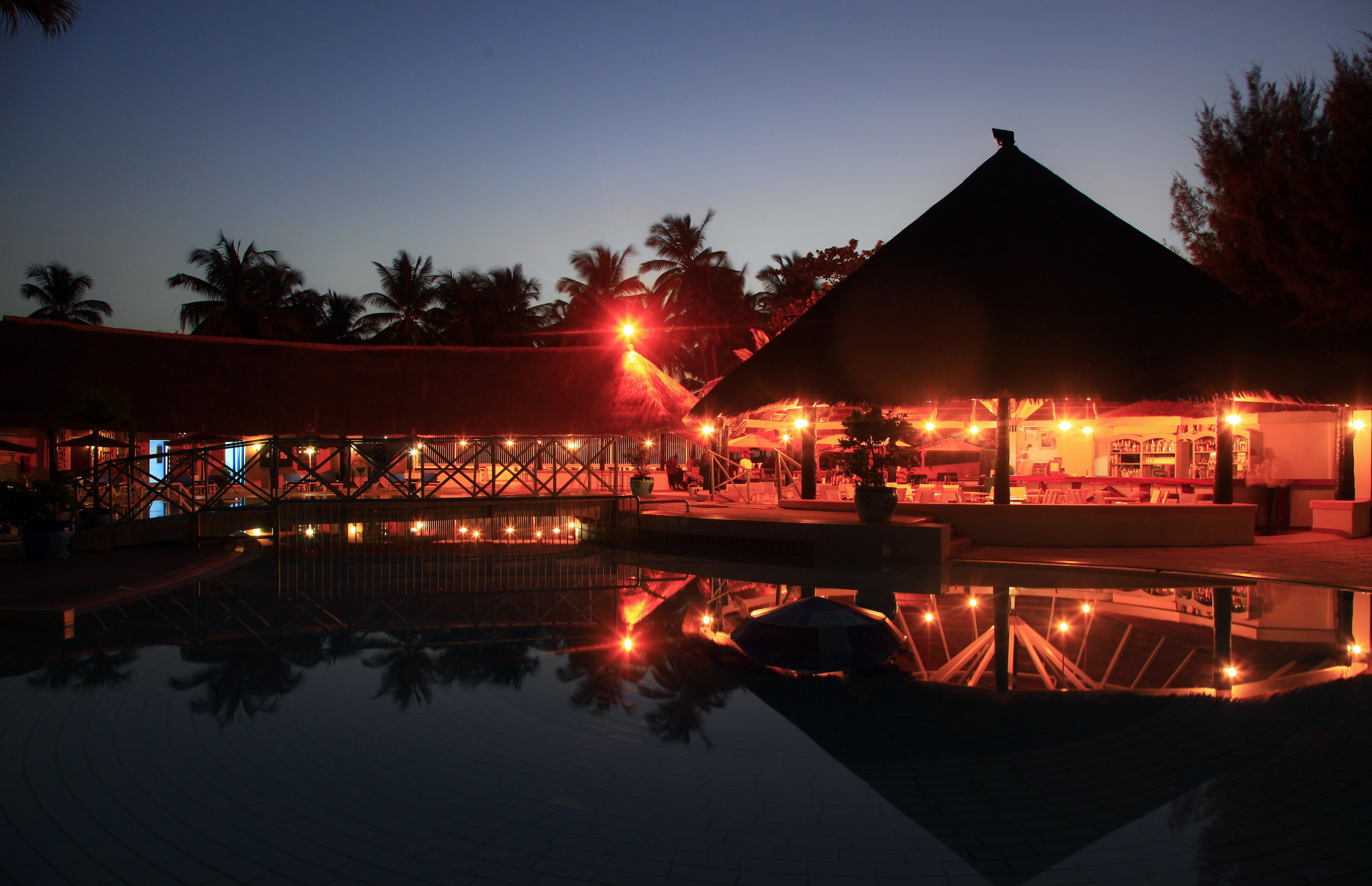 Et resort i skumringen med en pool der spejler lys, en bygning med stråtækt tag og palmer.