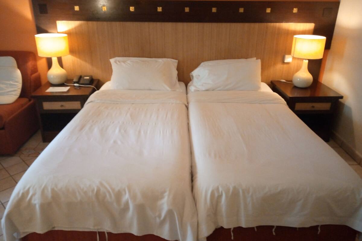 A hotel room with two single beds, each with a bedside table and lamp, and a wooden headboard.