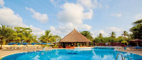 A large swimming pool with a thatched-roof pavilion, surrounded by palm trees and lounge chairs.