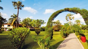 A pathway leading through a well-maintained garden with palm trees and a distinctive archway. - The Kairaba Beach Hotel (Serrekunda)