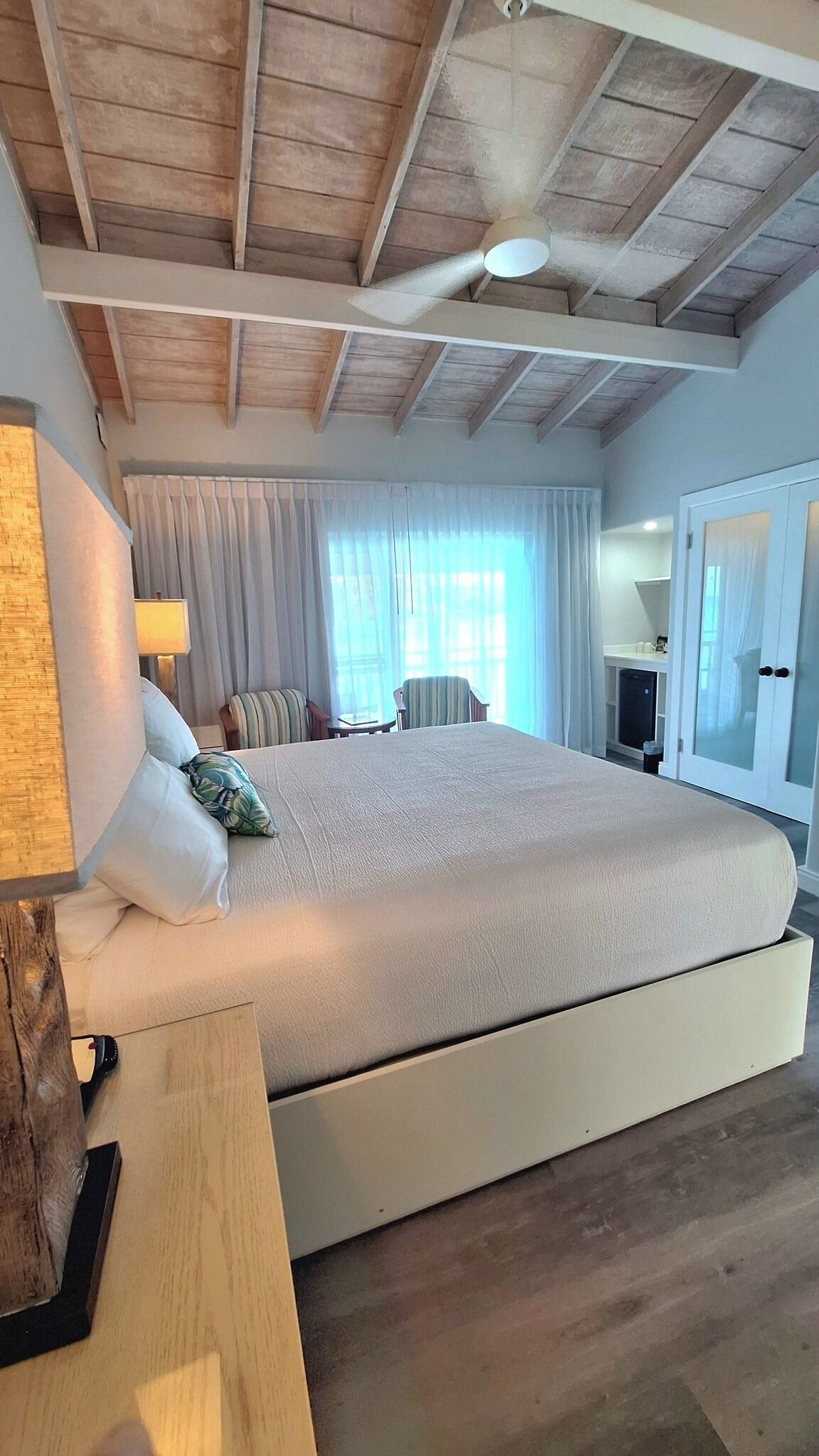 A spacious bedroom with a large bed, a bedside table, a lamp, a chair, a wardrobe, and a window with curtains.