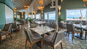 A spacious dining area with high ceilings and large windows overlooking the ocean. Tables are set with white tablecloths and glassware. - Pineapple Beach Club Antigua - Adults Only – All Inclusive (Willikies)