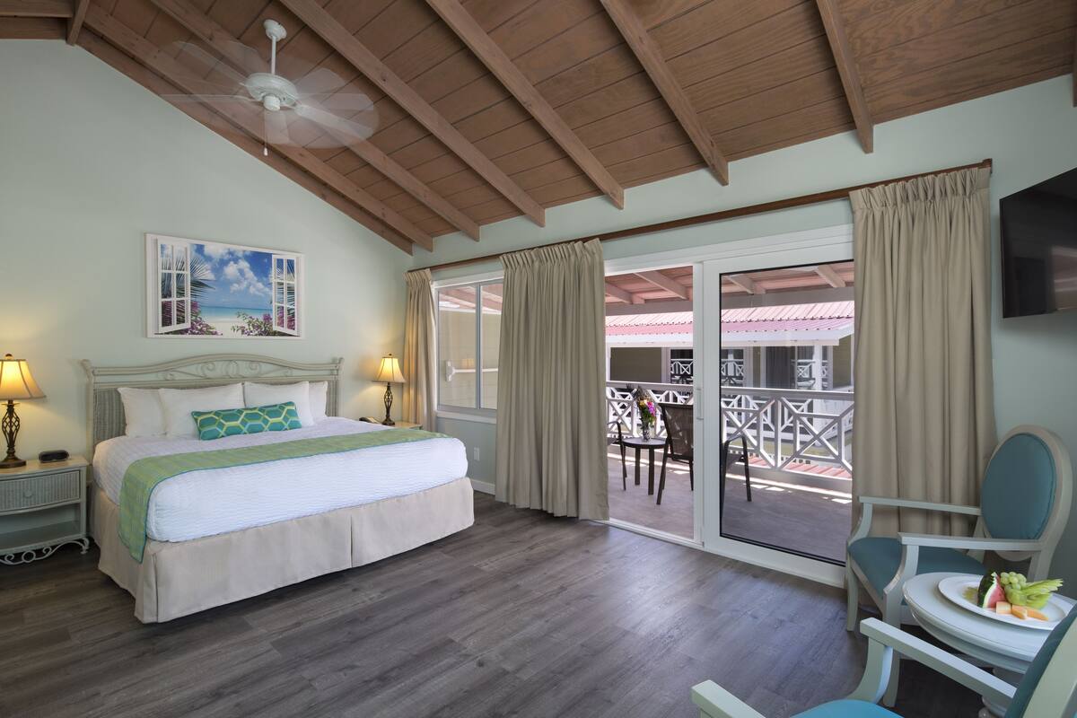 A hotel room with a bed, a balcony with a table and chairs, a ceiling fan, and a painting of a beach scene.