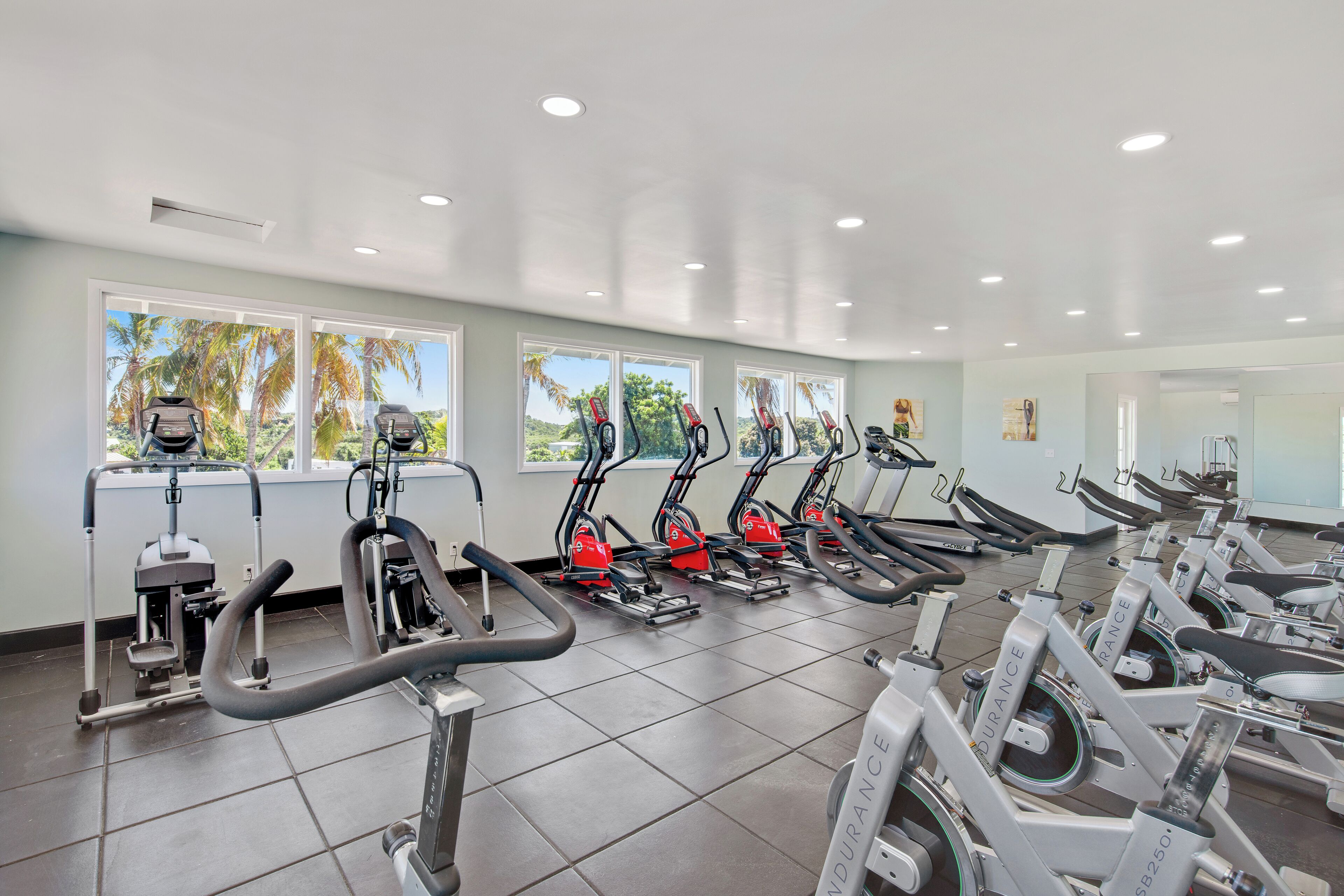 A gym with multiple cardio machines, large windows, and a tiled floor.