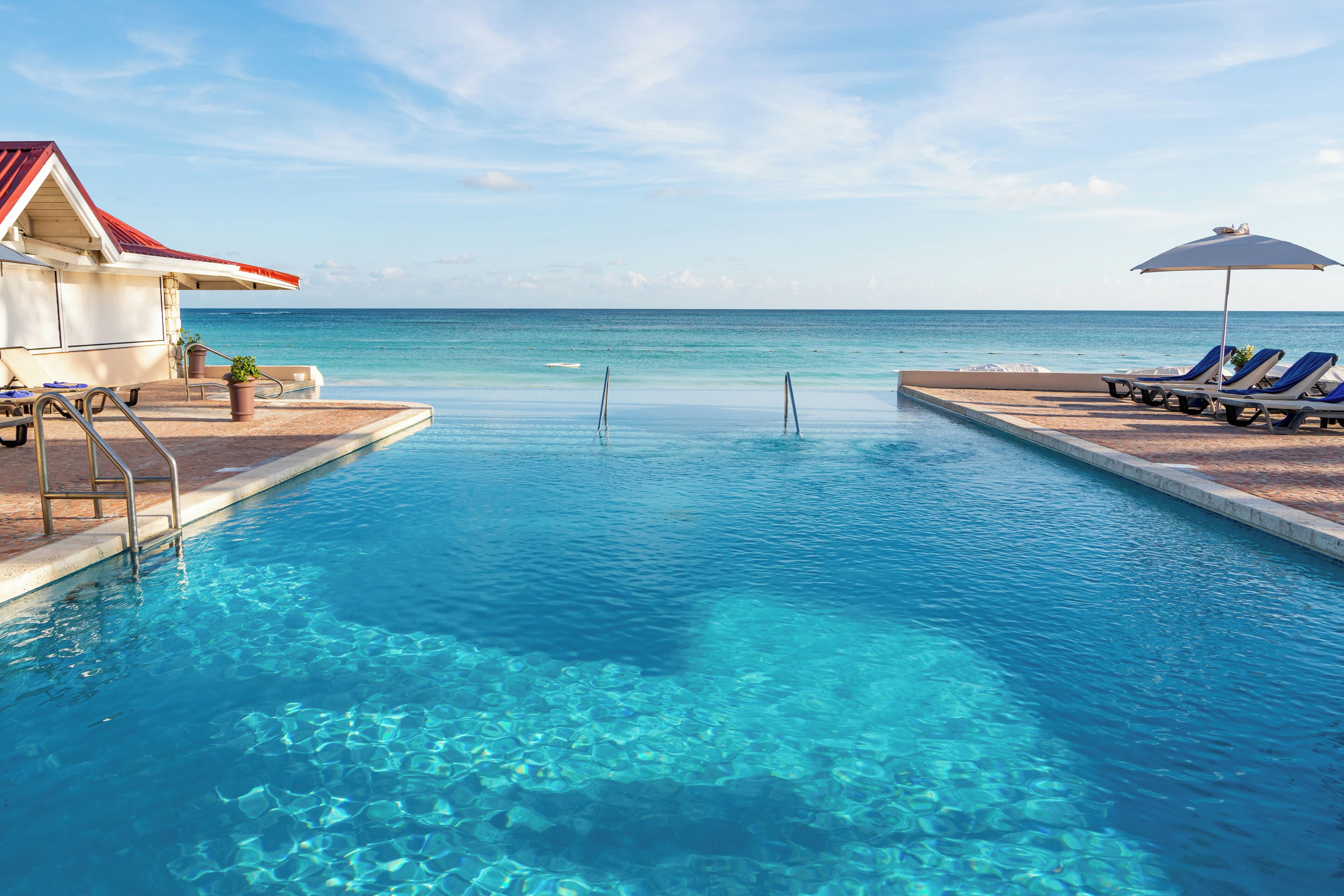 A clear pool with lounge chairs and umbrellas, a building with a red roof, and a view of the ocean.
