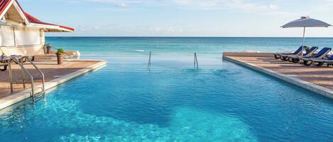 A clear pool with lounge chairs and umbrellas, a building with a red roof, and a view of the ocean.