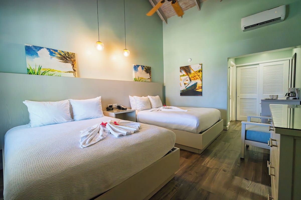 A hotel room with two beds, artwork on the walls, a desk with a chair, and an air conditioning unit.