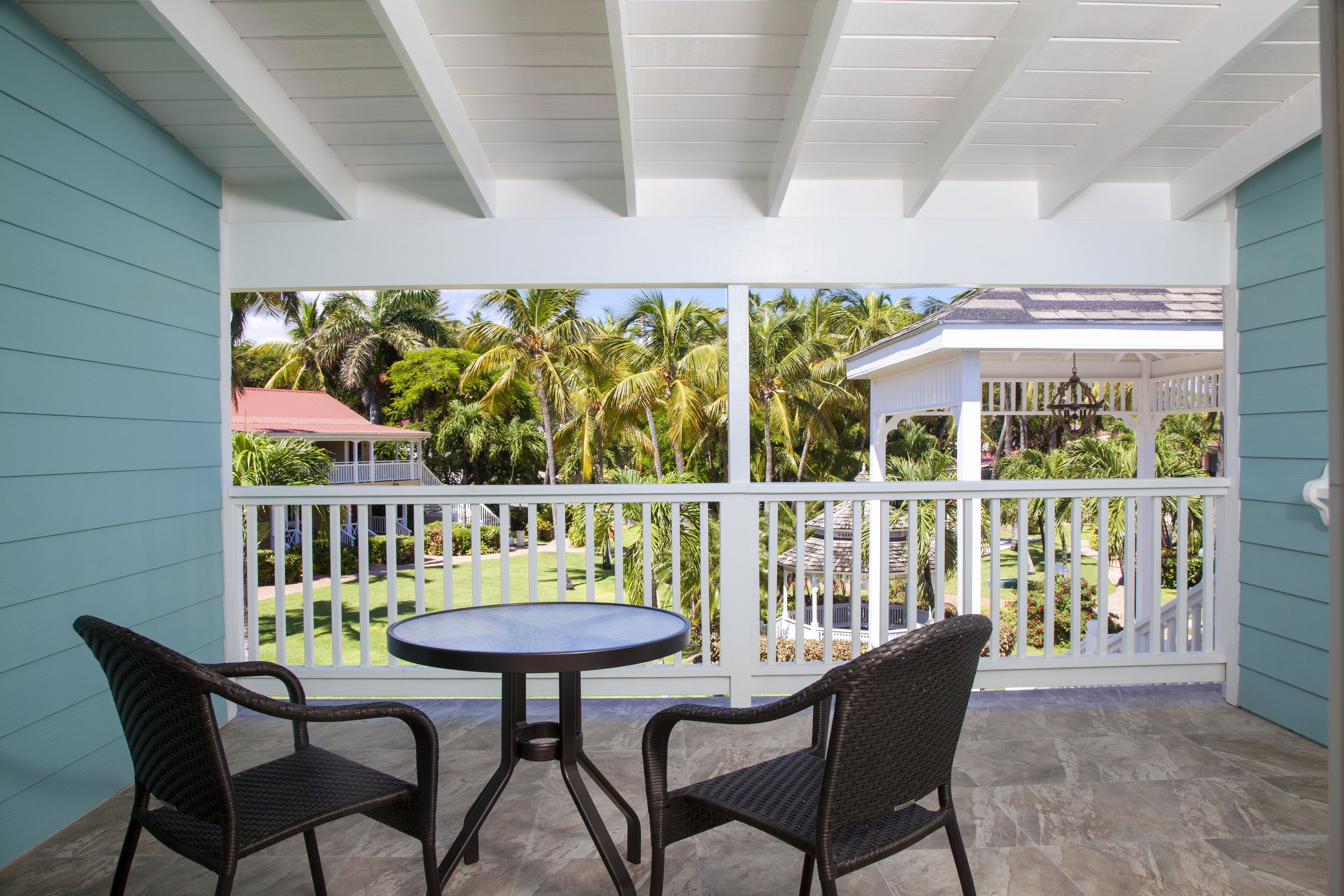 A covered patio with a round table and two wicker chairs, overlooking a garden with palm trees and a gazebo.