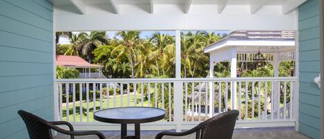 A covered patio with a round table and two wicker chairs, overlooking a garden with palm trees and a gazebo.