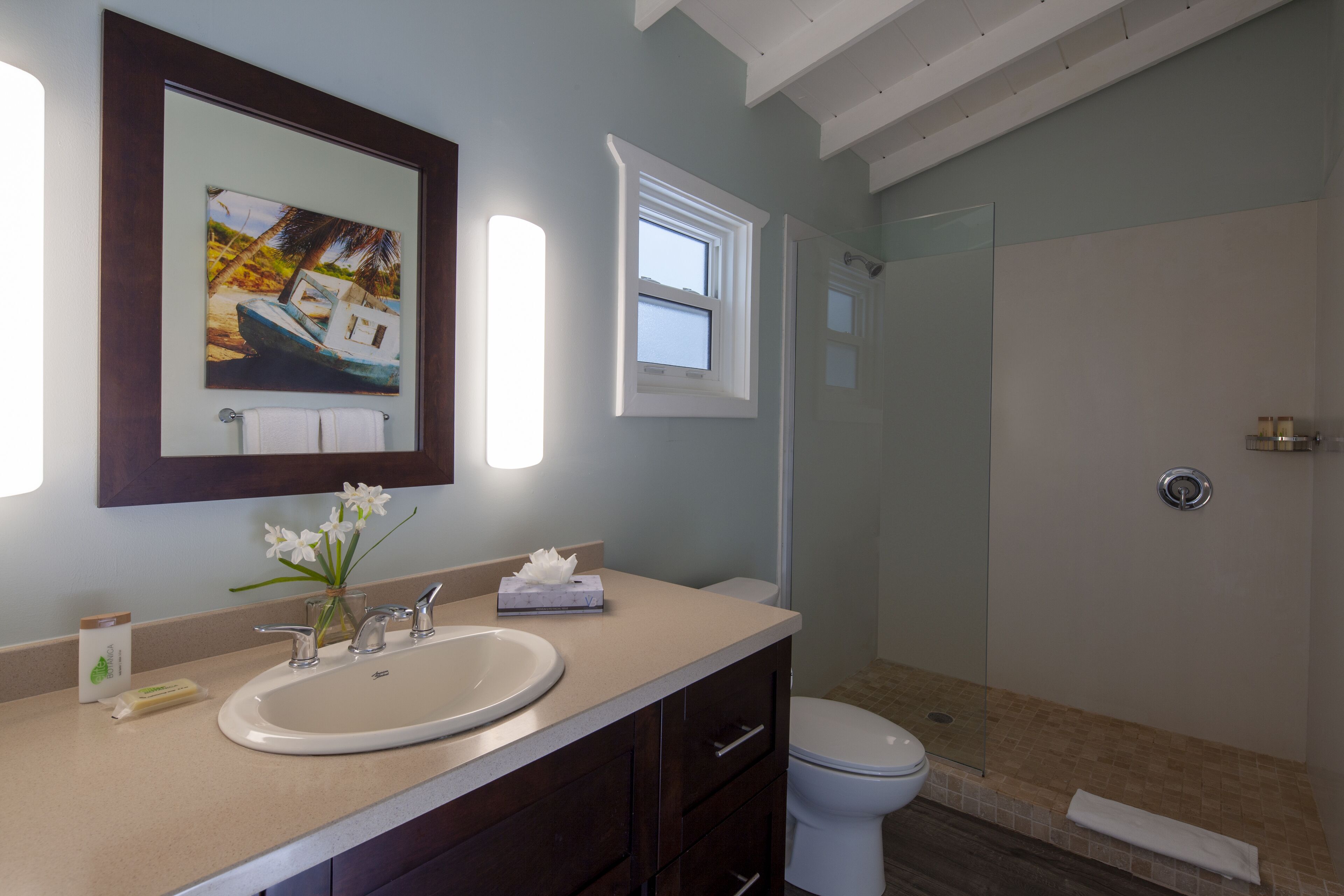 Une salle de bain équipée d’un lavabo, d’un miroir et de toilettes. Il y a une fenêtre au-dessus des toilettes et un espace douche avec une porte en verre.