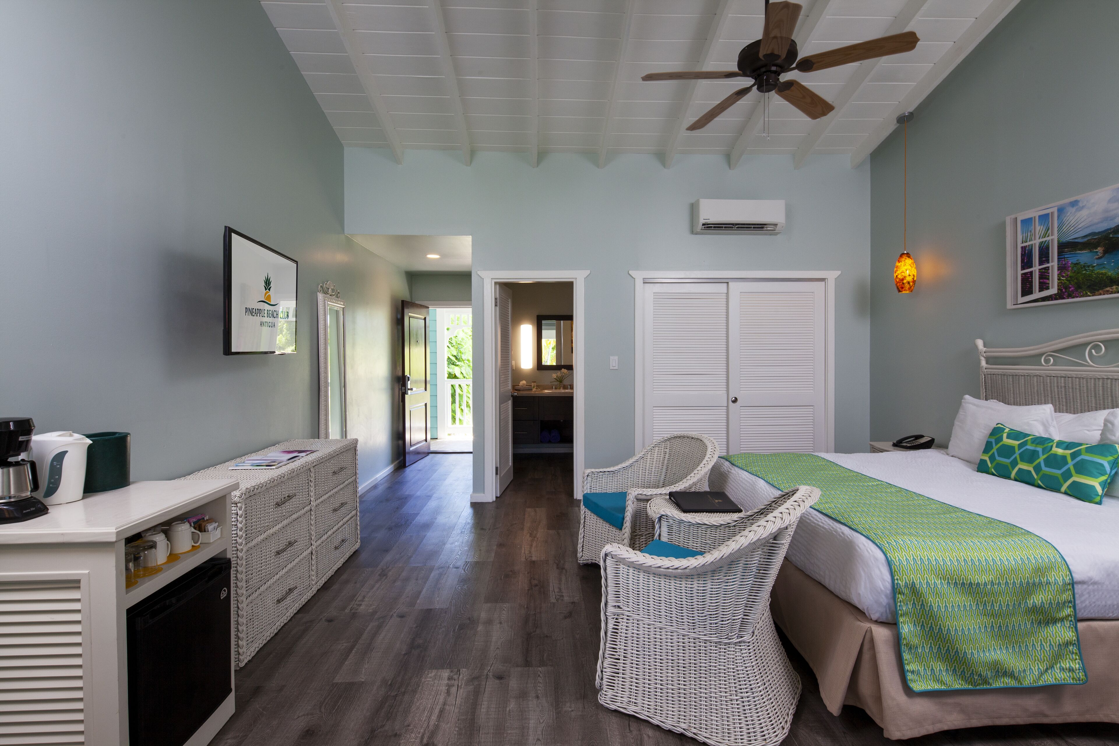 A hotel room with a bed, two wicker chairs, a desk with a coffee maker, and a ceiling fan.