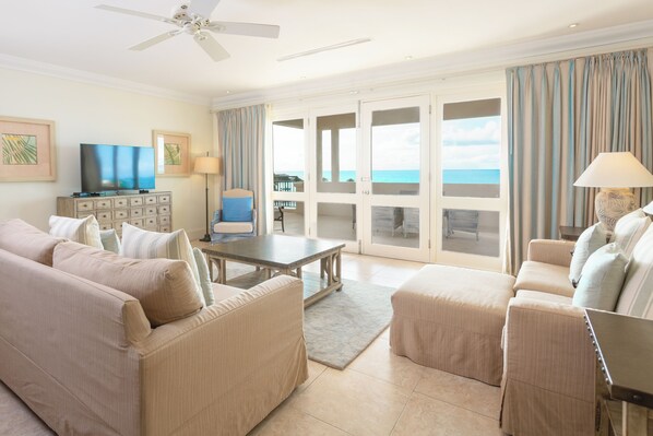 A spacious living room with a large sofa set, a coffee table, and a TV on a cabinet. There is a balcony with a view of the ocean. - Blue Waters Resort & Spa (St. John's)