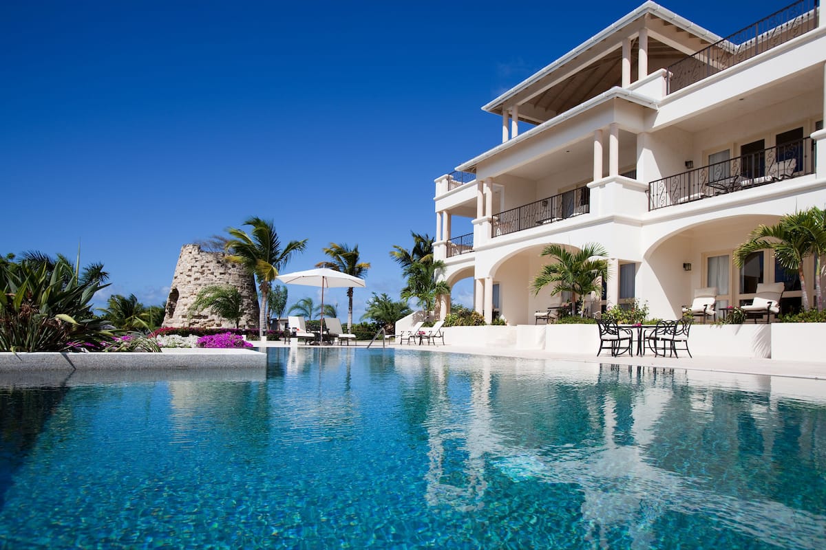 A large, clear swimming pool with lounge chairs and umbrellas, surrounded by palm trees and a white building with balconies.