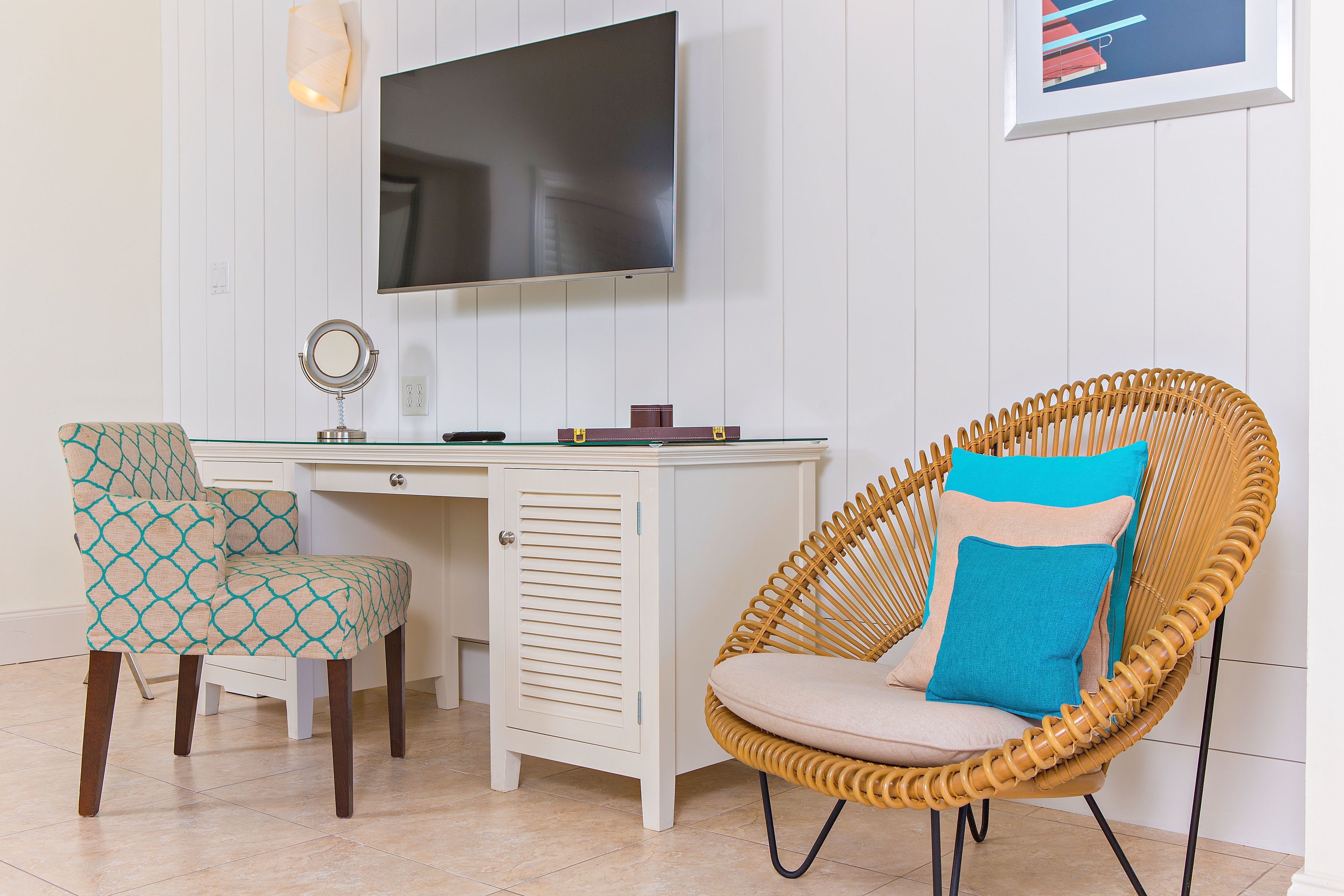 A room with a white desk, a television mounted on the wall, a wicker chair with a cushion, and a patterned chair.