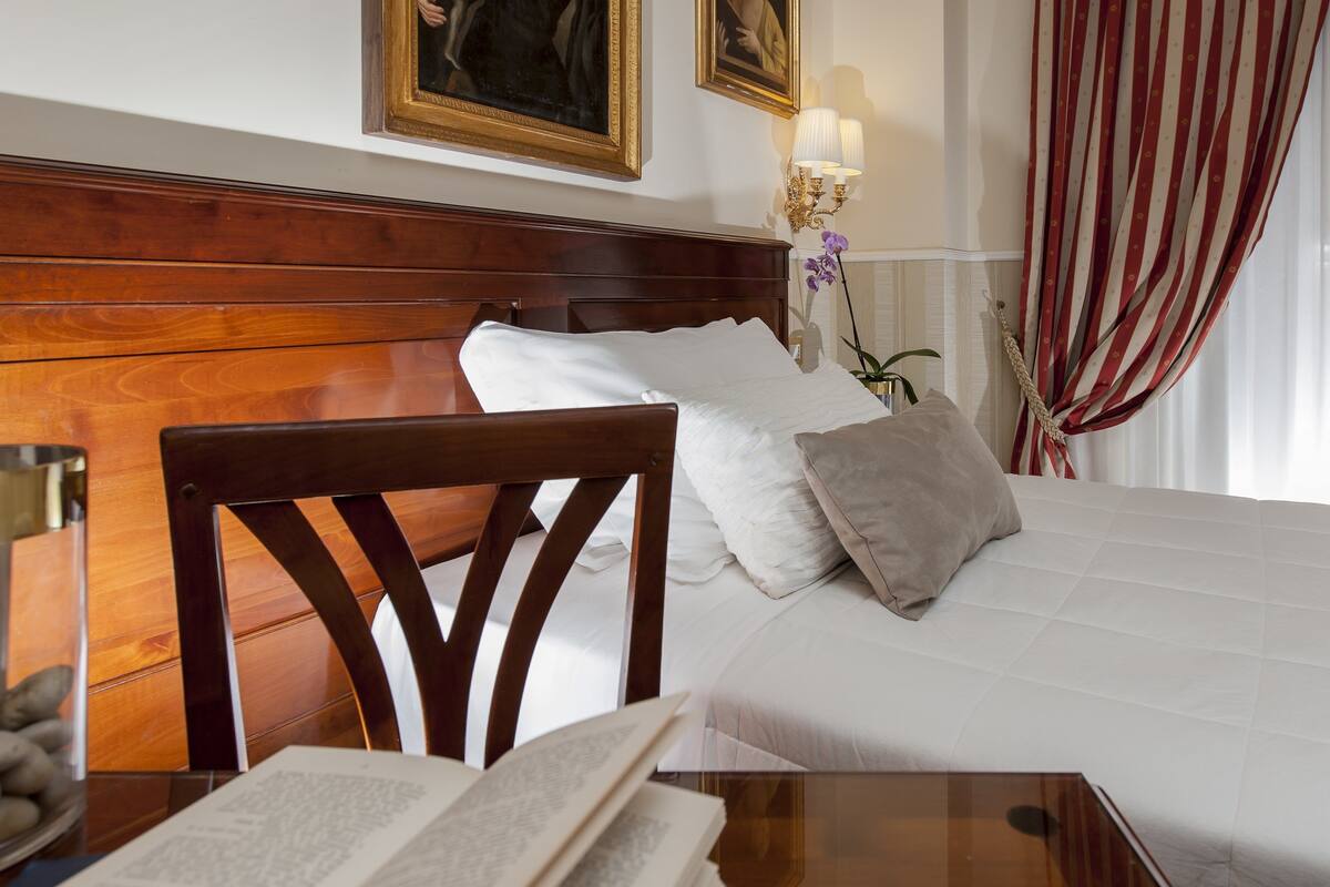 A hotel room with a wooden headboard, a bed with white linens, a nightstand with a book, and a wall-mounted lamp.