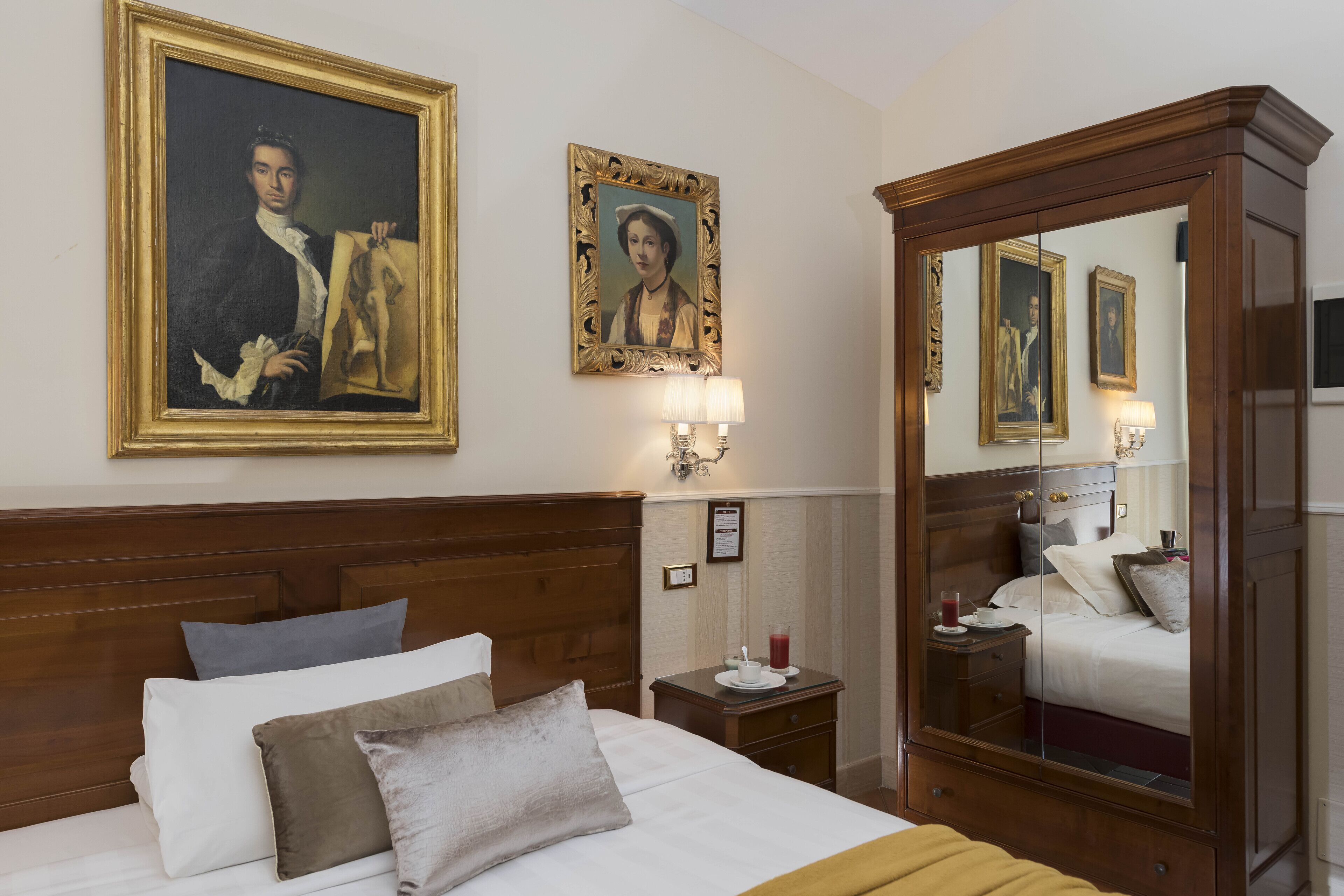 Habitación de hotel con una cama grande, cabecera de madera, mesita de noche, espejo y dos retratos enmarcados en la pared.
