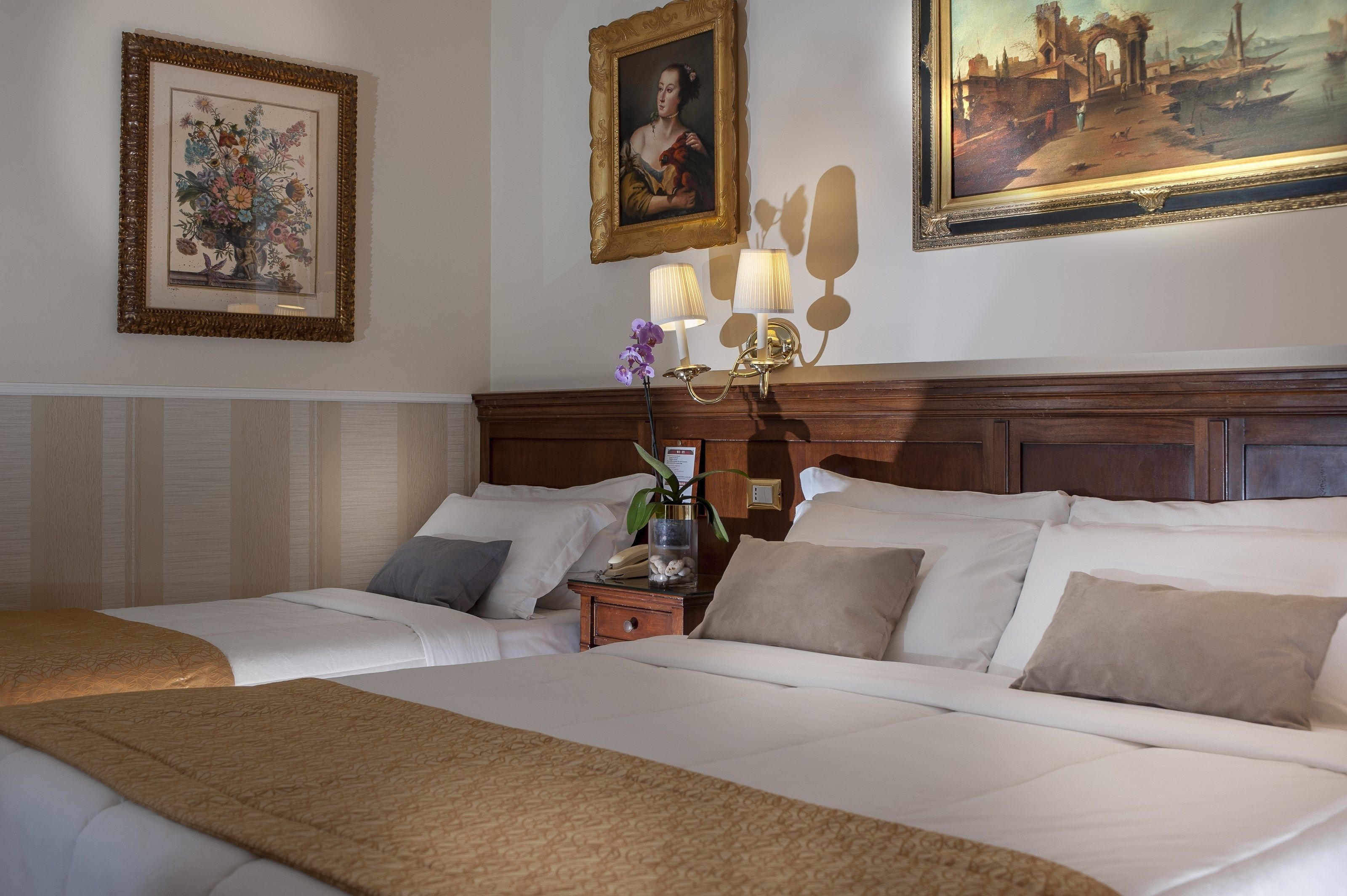 A hotel room with two beds, a nightstand with a lamp, and framed artwork on the walls.