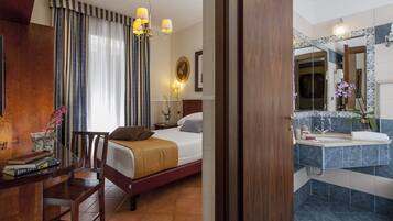 A hotel room with a bed, bedside table with books, a chair, a desk with a vase of flowers, a bathroom with a sink, mirror, and blue tiles.