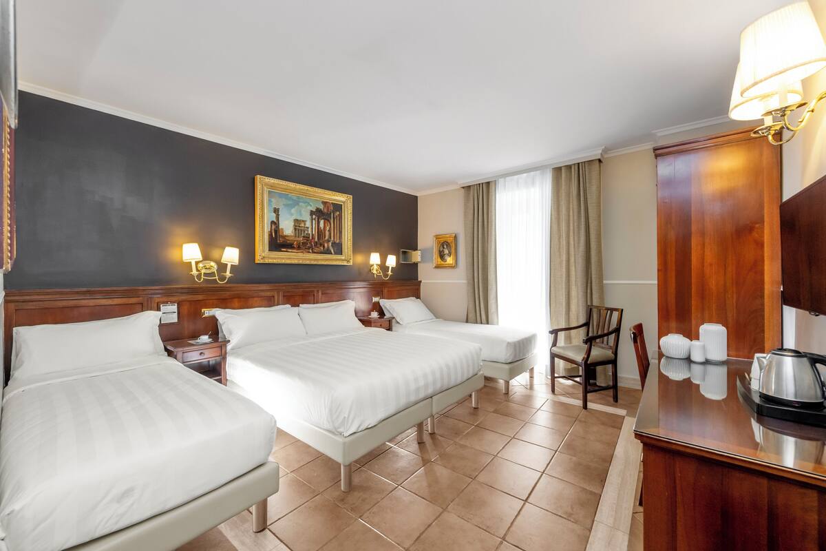 A hotel room with two beds, a wooden headboard, a desk with a lamp, a chair, and a painting on the wall.