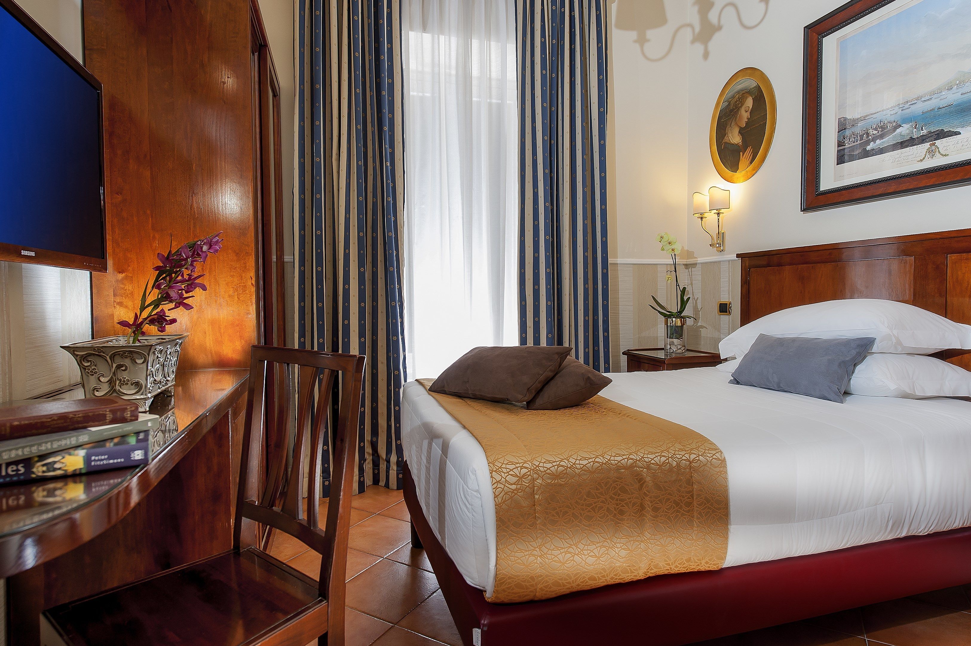 A hotel room with a bed, a desk with books, a chair, a TV, curtains, a framed picture, and a nightstand with a lamp.