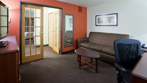A hotel room with a wooden desk, a small round table, a patterned sofa, a blue office chair, and a sliding door leading to a bathroom. - AmericInn by Wyndham Grafton (Grafton)