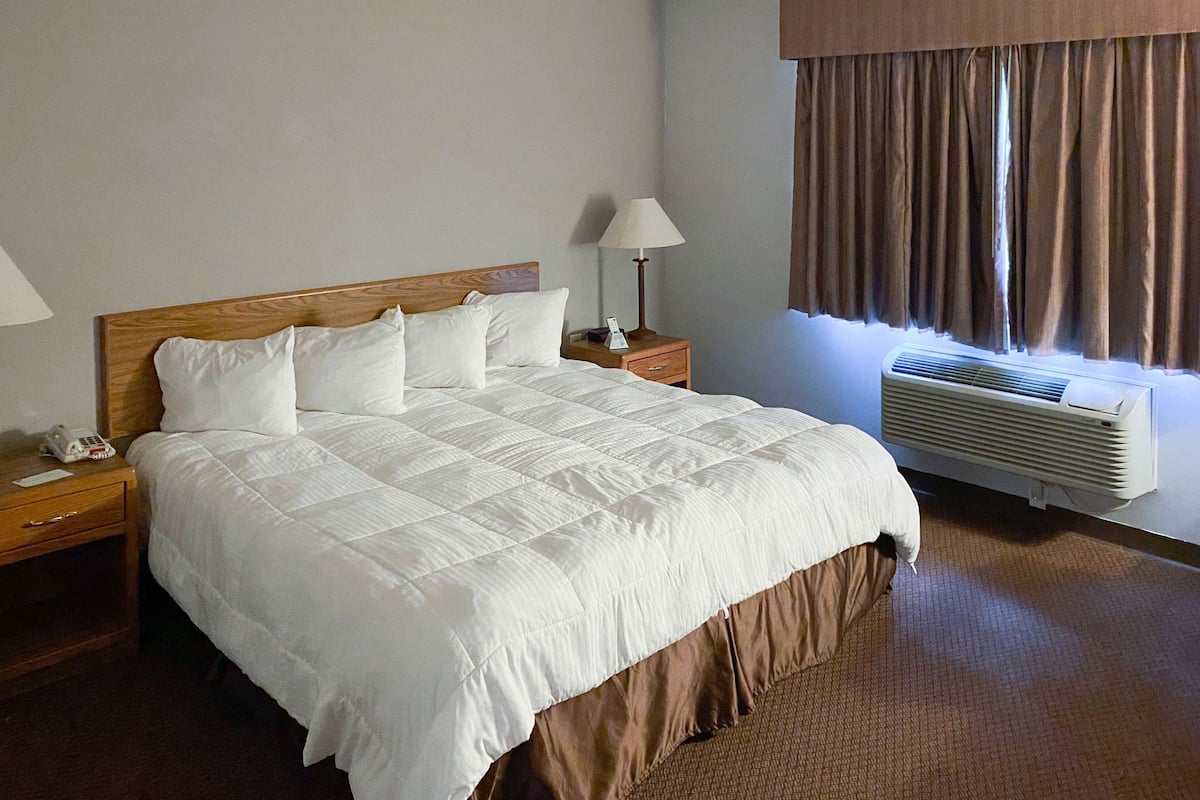 A hotel room with a large bed, a bedside table with a lamp, a nightstand, and an air conditioning unit.