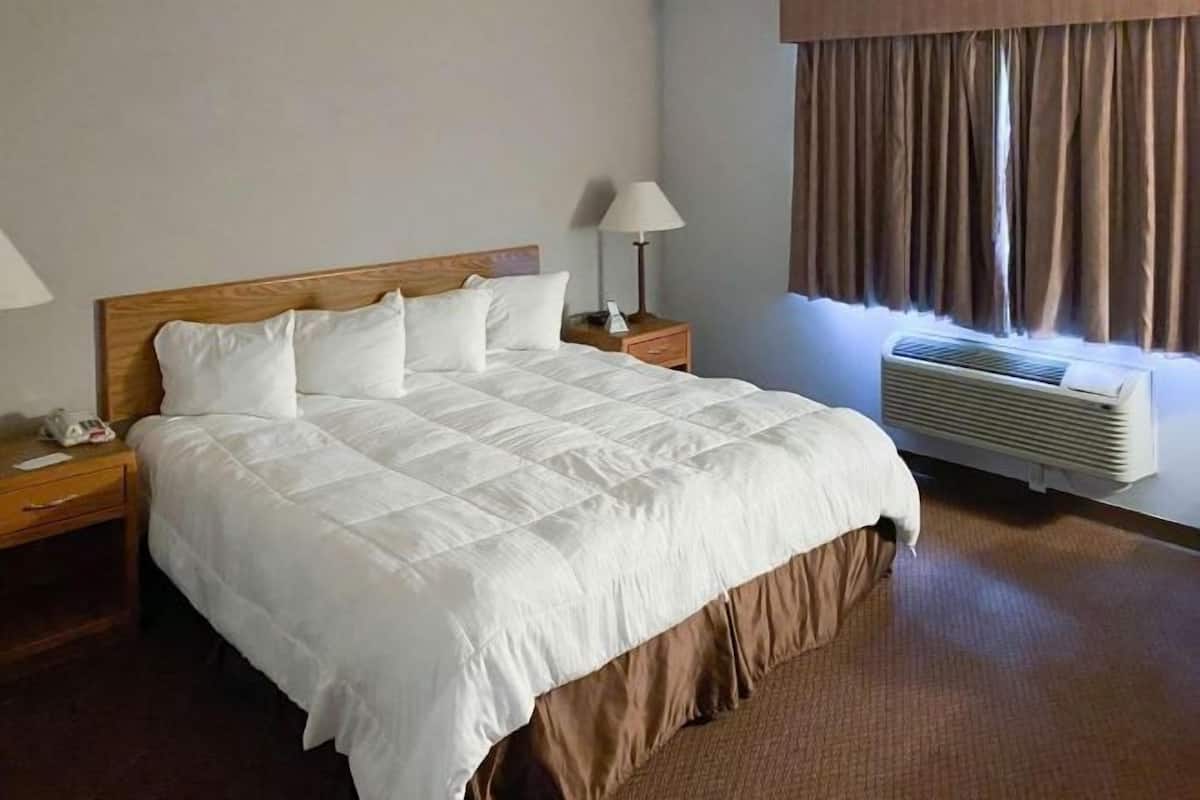 A hotel room with a large bed, a bedside table with a lamp, a window with curtains, and a wall-mounted air conditioner.