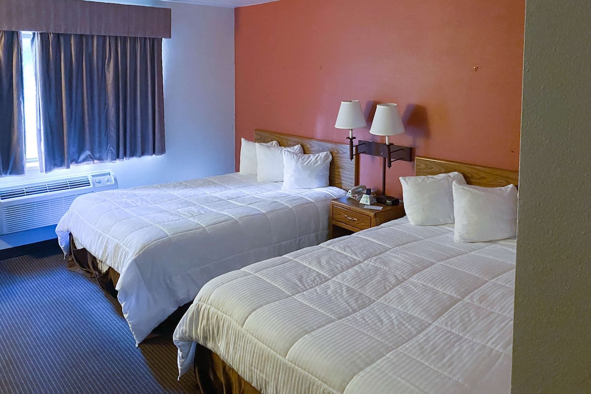 A hotel room with two beds, a nightstand with lamps, a window with curtains, and an air conditioning unit.