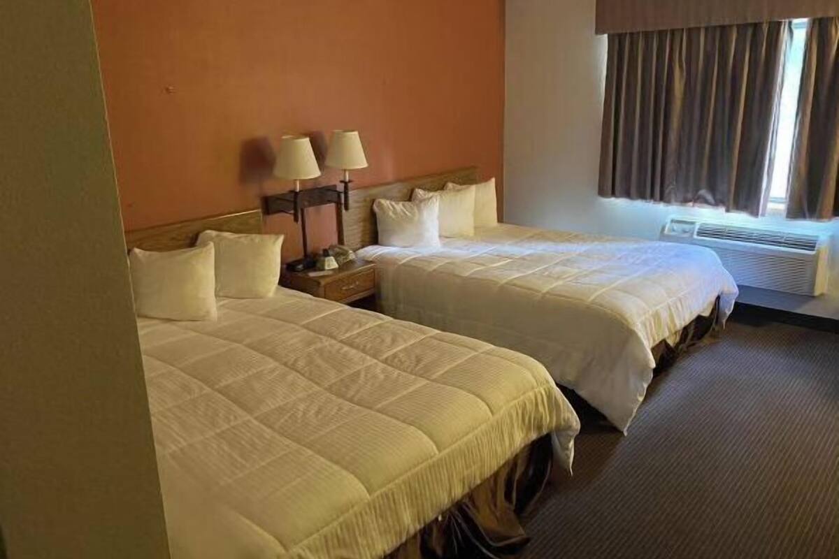 A hotel room with two beds, a nightstand, lamps, a window with curtains, and an air conditioning unit.