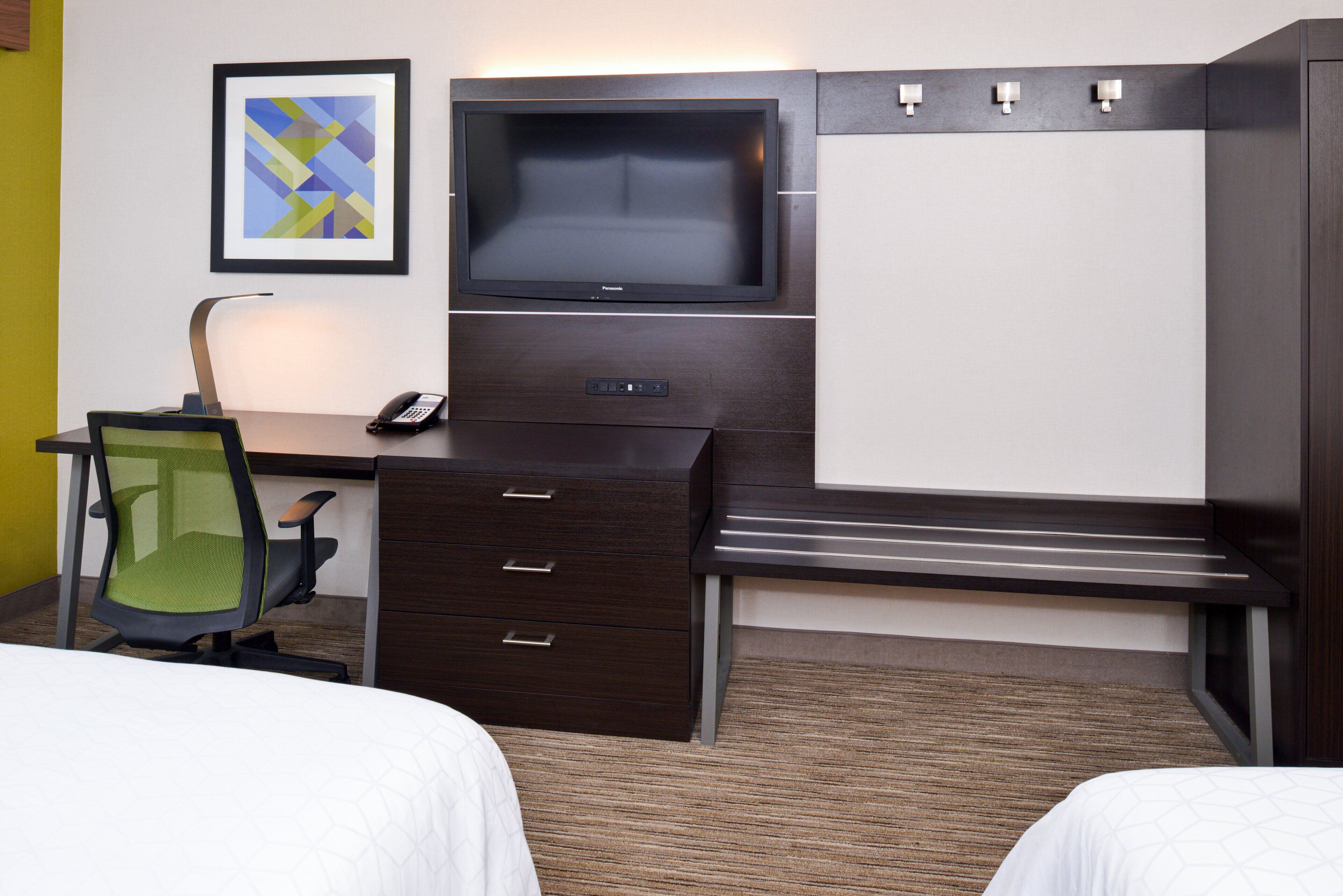A hotel room with a dark wood desk, a flat-screen TV, a green mesh office chair, a bedside table, and a bed with white bedding.