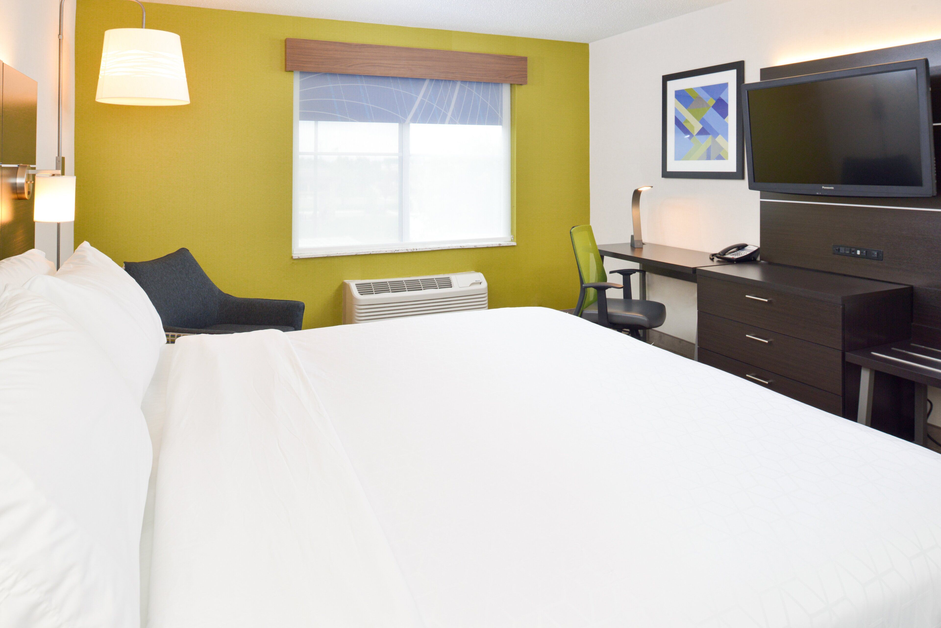 Quarto de hotel com cama, secretária, cadeira, televisão e janela com persianas.