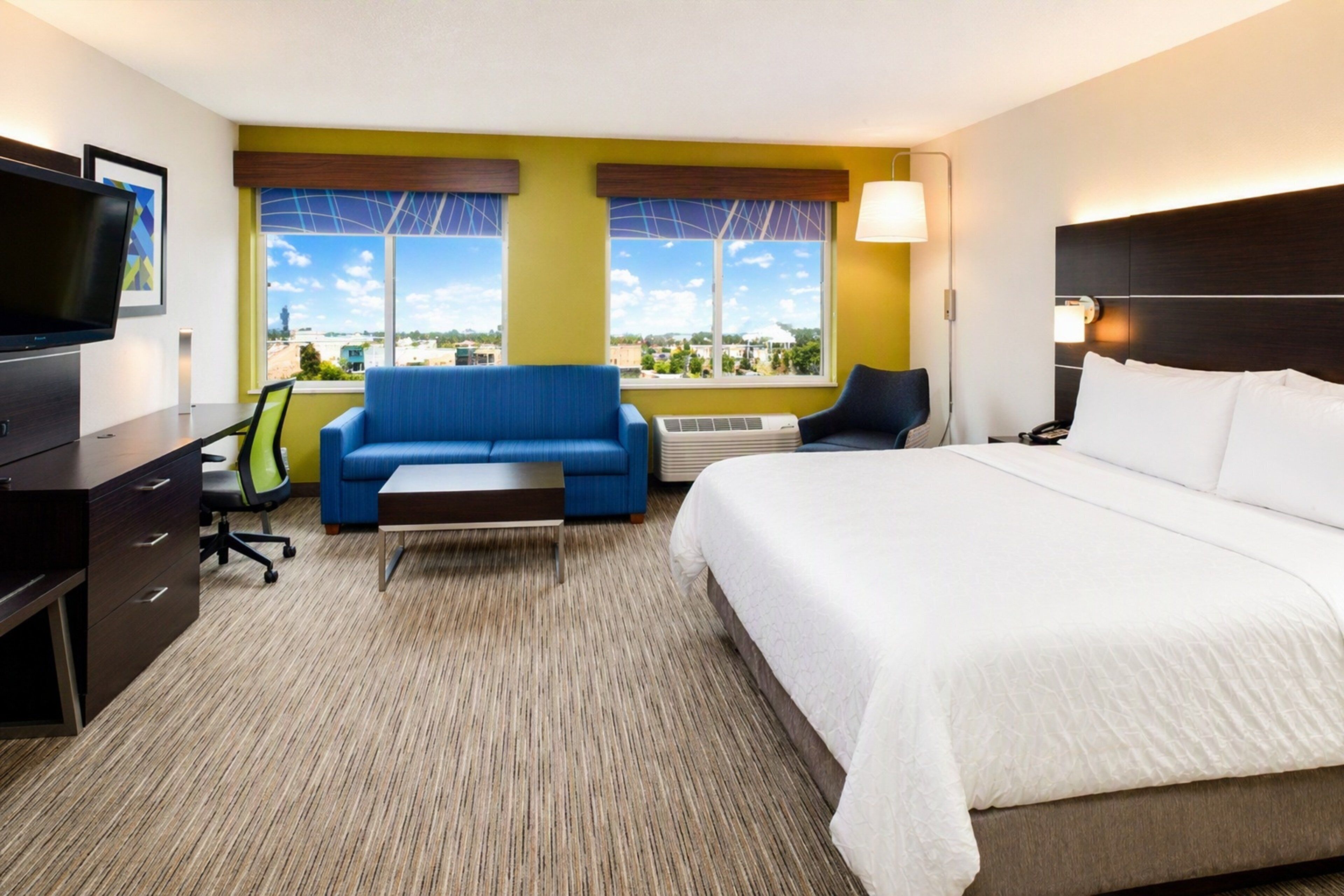 A hotel room with a bed, a desk, a chair, a sofa, and large windows with a view.