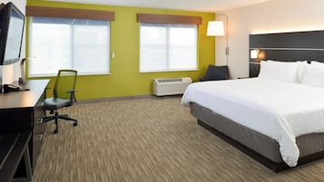 Una habitación de hotel con cama, escritorio con silla, dos ventanas con persianas, aire acondicionado y televisor sobre un mueble.