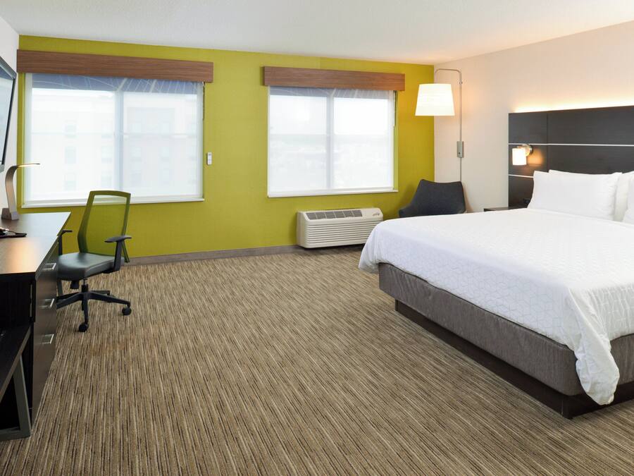 Une chambre d’hôtel comprenant un lit, un bureau avec une chaise, deux fenêtres équipées de stores, un climatiseur et un téléviseur posé sur un meuble.