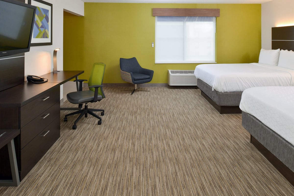 Une chambre d’hôtel avec un lit, un bureau avec une télévision, une chaise et une fenêtre avec des stores.