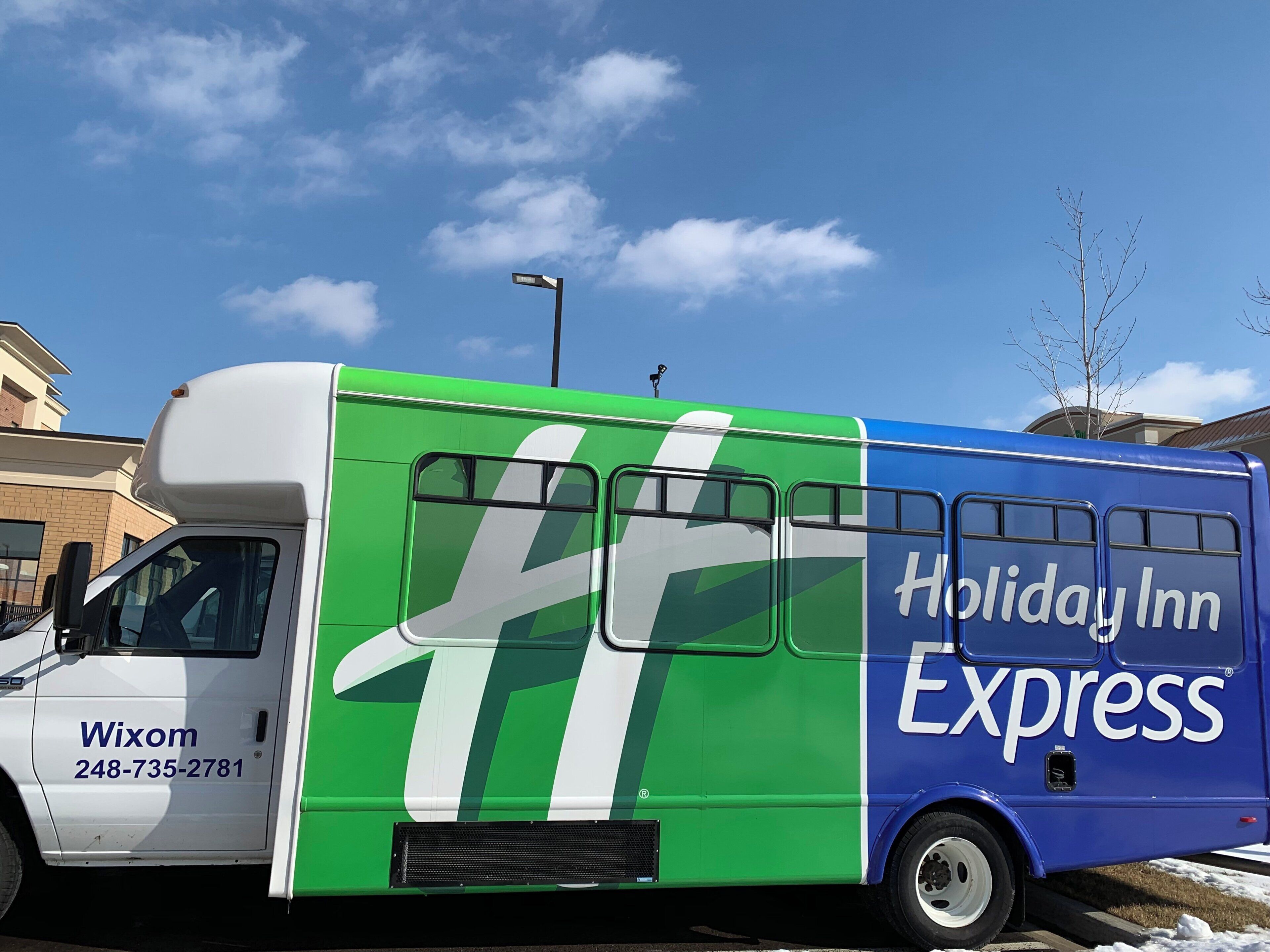 Um ônibus Holiday Inn Express com uma grande logo verde e branca, o nome 'Holiday Inn Express' na lateral, e o número de telefone 'Wixom 248-735-2781' na frente.