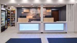 Een receptie van een hotel met een modern design, met een centraal gelegen bureau met verlichte panelen, een koelkast gevuld met snacks en een wandbord met de tekst Holiday Inn Express & Suites.