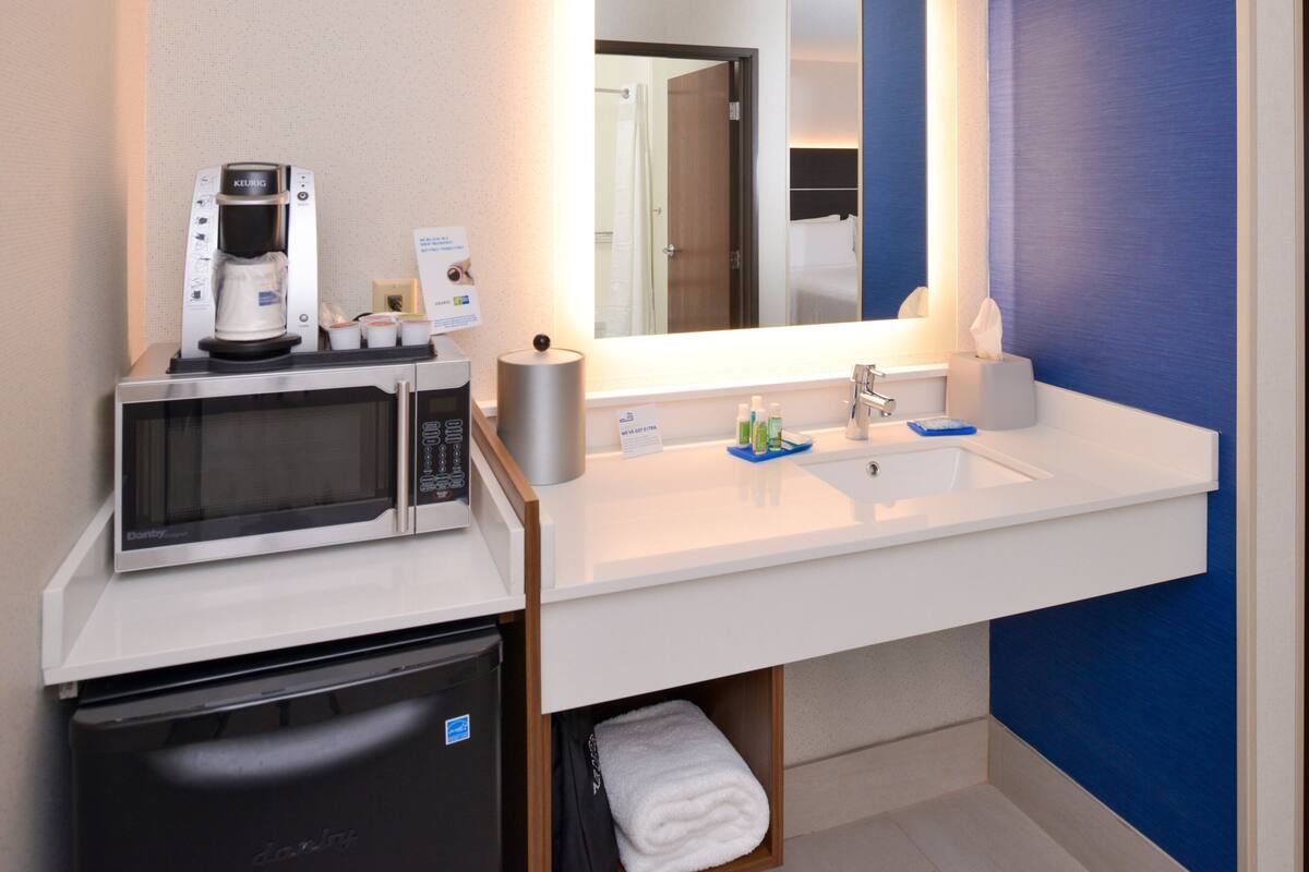 Une salle de bain d’hôtel avec un lavabo blanc, un miroir, une machine à café, un four à micro-ondes et un petit réfrigérateur.