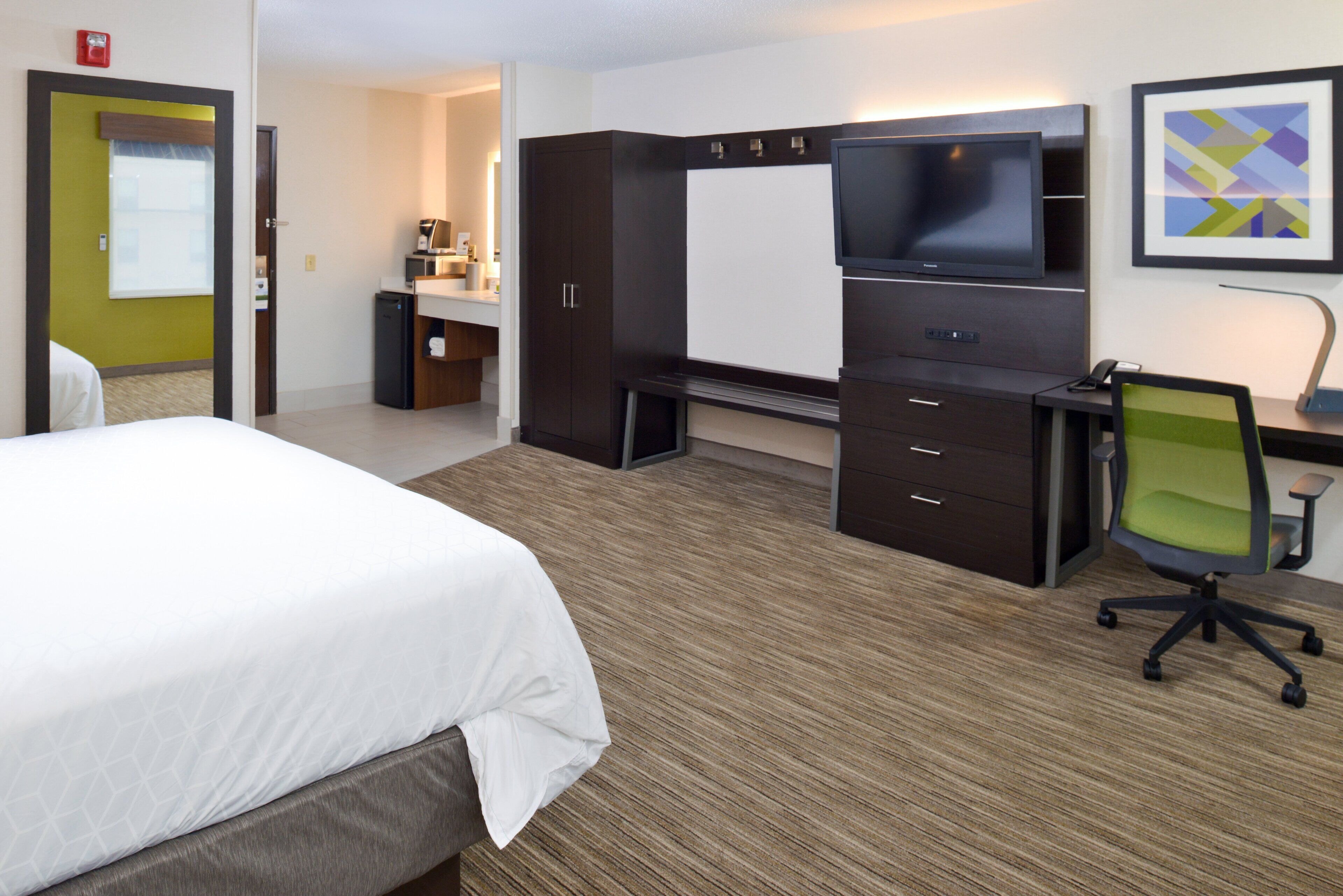 A hotel room with a bed, a desk with a chair, a TV, and a kitchenette.