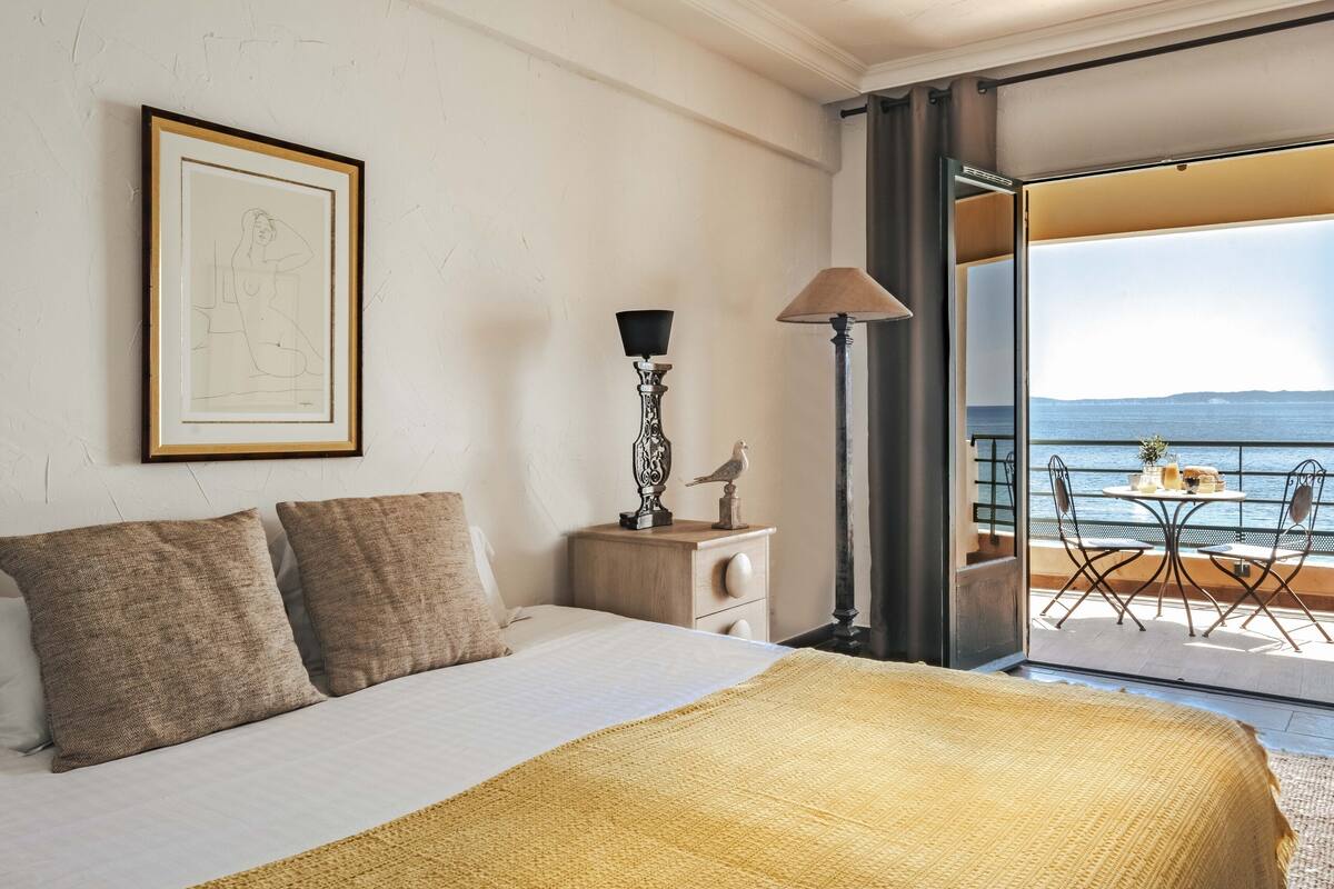A hotel room with a bed, a framed artwork, a nightstand with a lamp, and a balcony with a table and chairs.