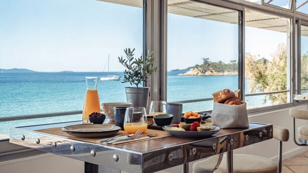 Una mesa de desayuno con vista al mar, una planta en maceta y una cesta de cruasanes.