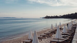 Una playa con hileras de sillas de madera y sombrillas blancas, un mar tranquilo y una costa distante con árboles.
