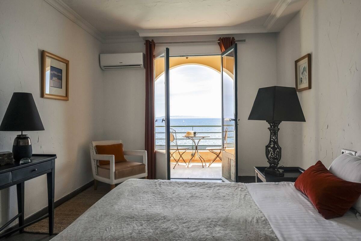 A hotel room with a bed, a chair, a table, and a view of the beach through an open door.