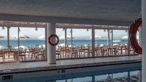 Zona de piscina interior con vista al mar, mesas y sillas de madera, y un flotador colgado en la pared.