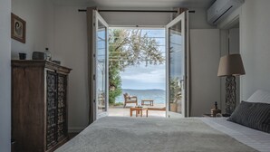 Un dormitorio con una cama grande, un armario de madera, una mesita de noche, una lámpara y vistas al mar a través de una puerta abierta.
